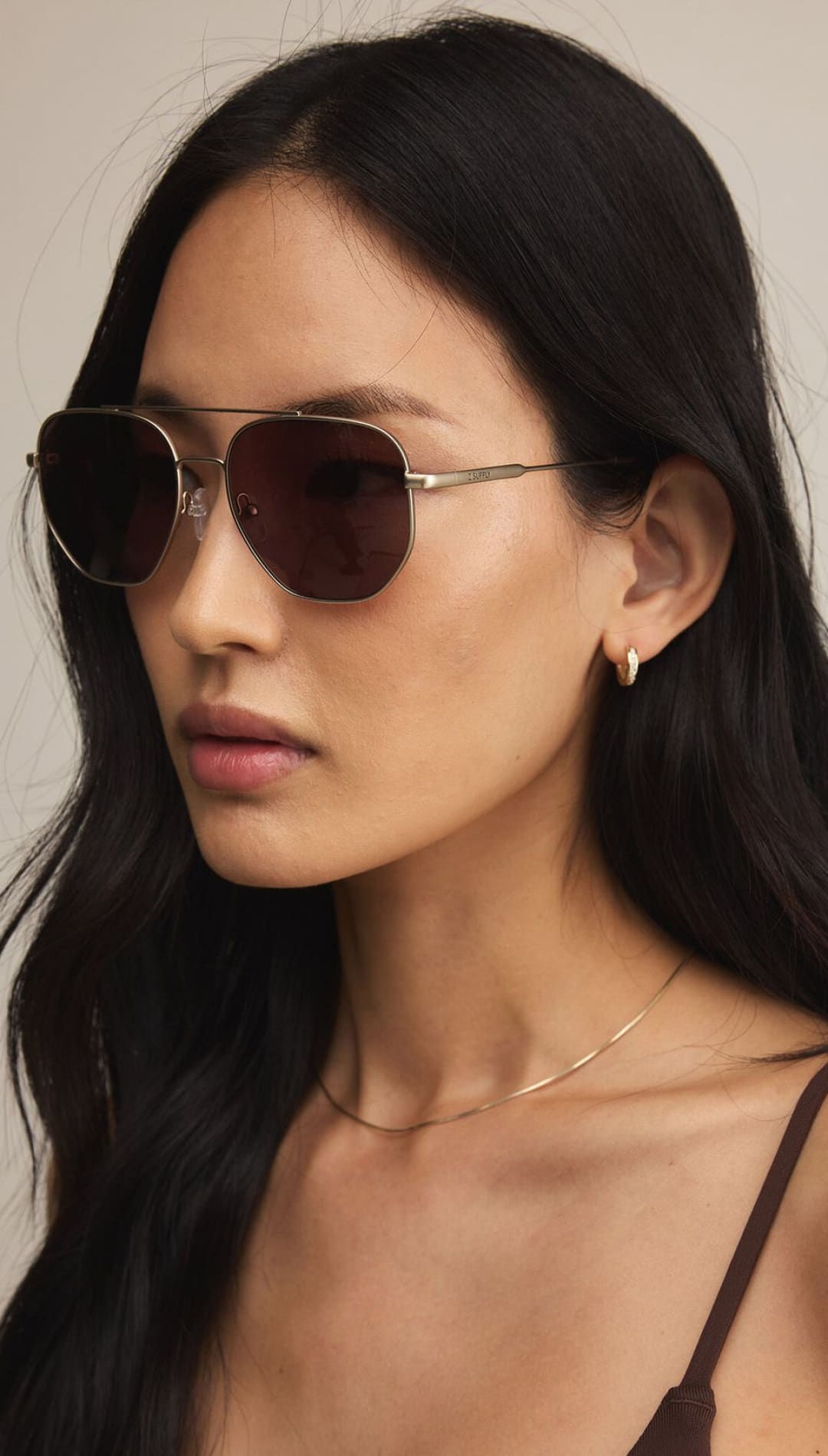 A woman with long dark hair wearing aviator sunglasses, a delicate gold chain necklace, and a brown top.