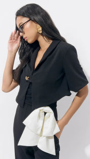 A woman wearing a black cropped blazer with a bow detail and dark trousers.