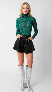 Green and black long-sleeve turtleneck top with black faux leather shorts and silver knee-high boots.