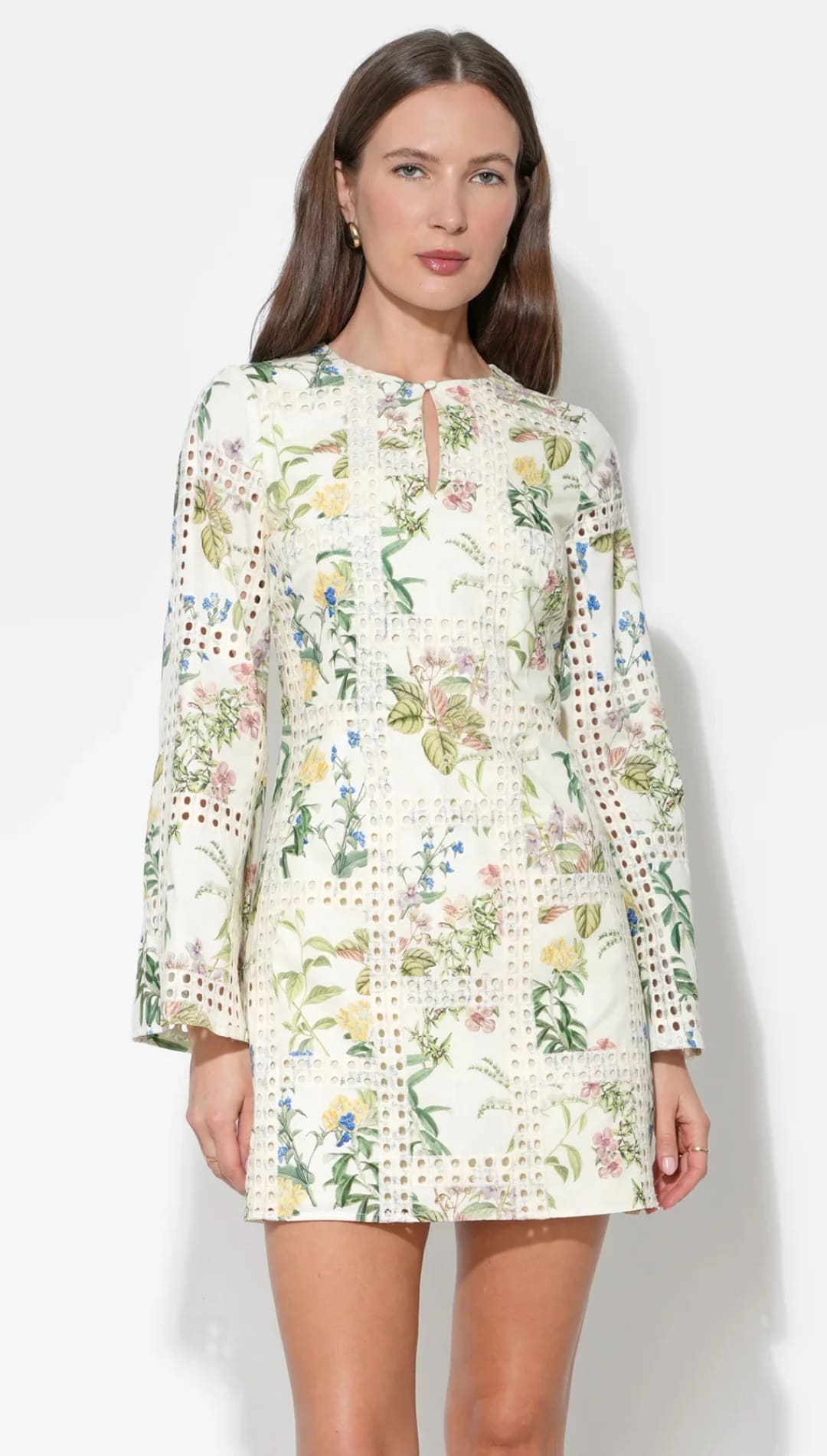 A woman wears a cream-colored, long-sleeved dress featuring an intricate floral pattern with green leaves and soft pastel blossoms, accented by delicate lace detailing along the cuffs and hem.