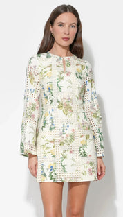 A woman wears a cream-colored, long-sleeved dress featuring an intricate floral pattern with green leaves and soft pastel blossoms, accented by delicate lace detailing along the cuffs and hem.