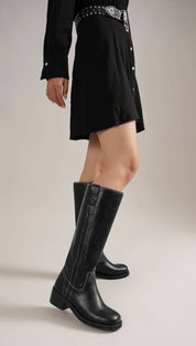Woman wearing a black collared shirt dress with a studded belt and black knee-high boots with white stitching.