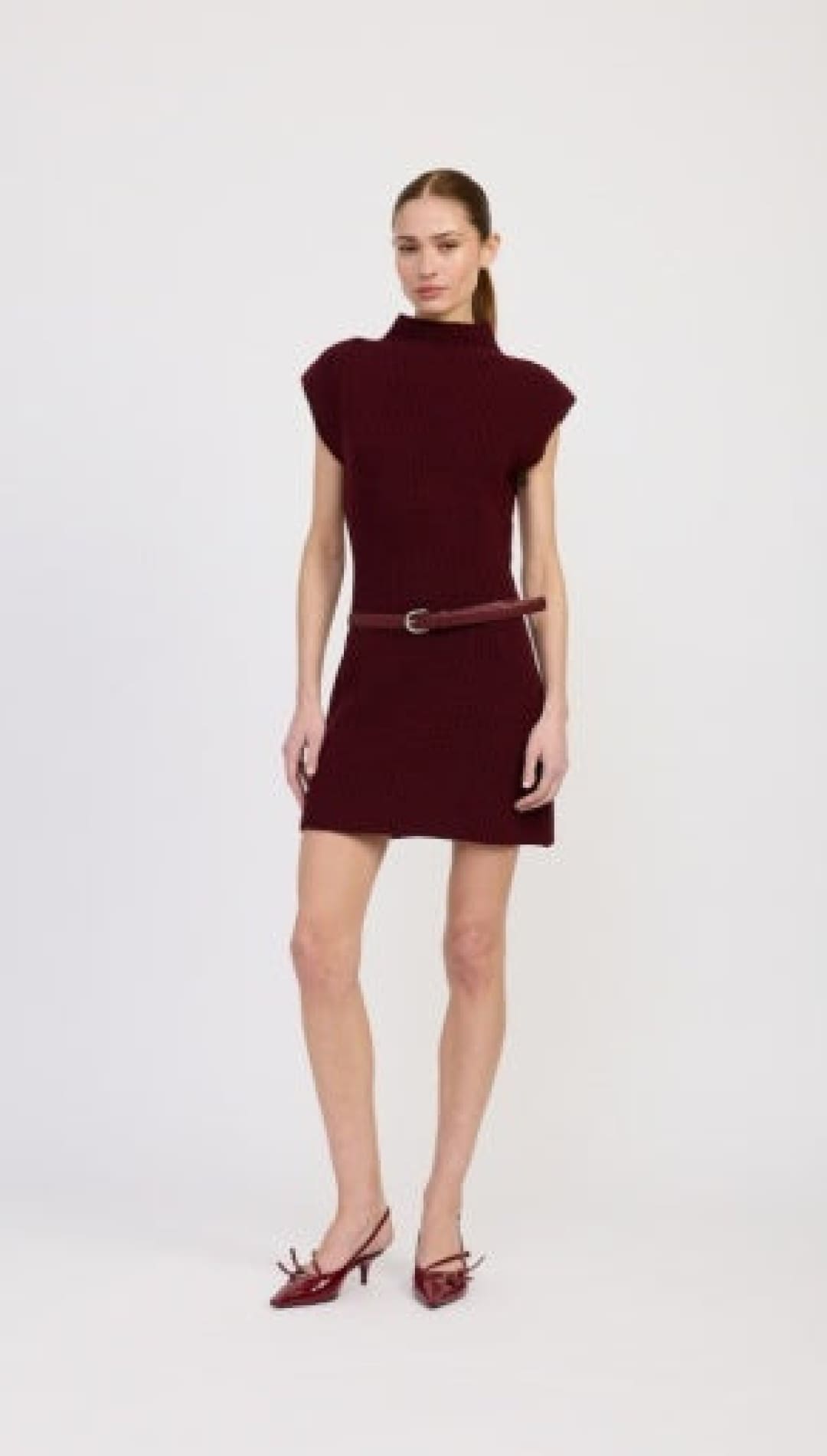 A model wearing a short-sleeved, high-necked, burgundy mini dress with a thin matching belt and pointed-toe kitten heels with bows.