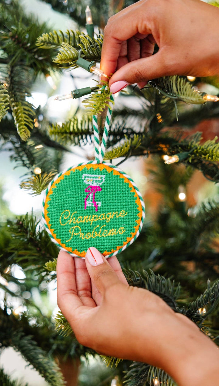 Green cross-stitch ornament with champagne glass and ’champagne problems’ text, wrapped in twine.