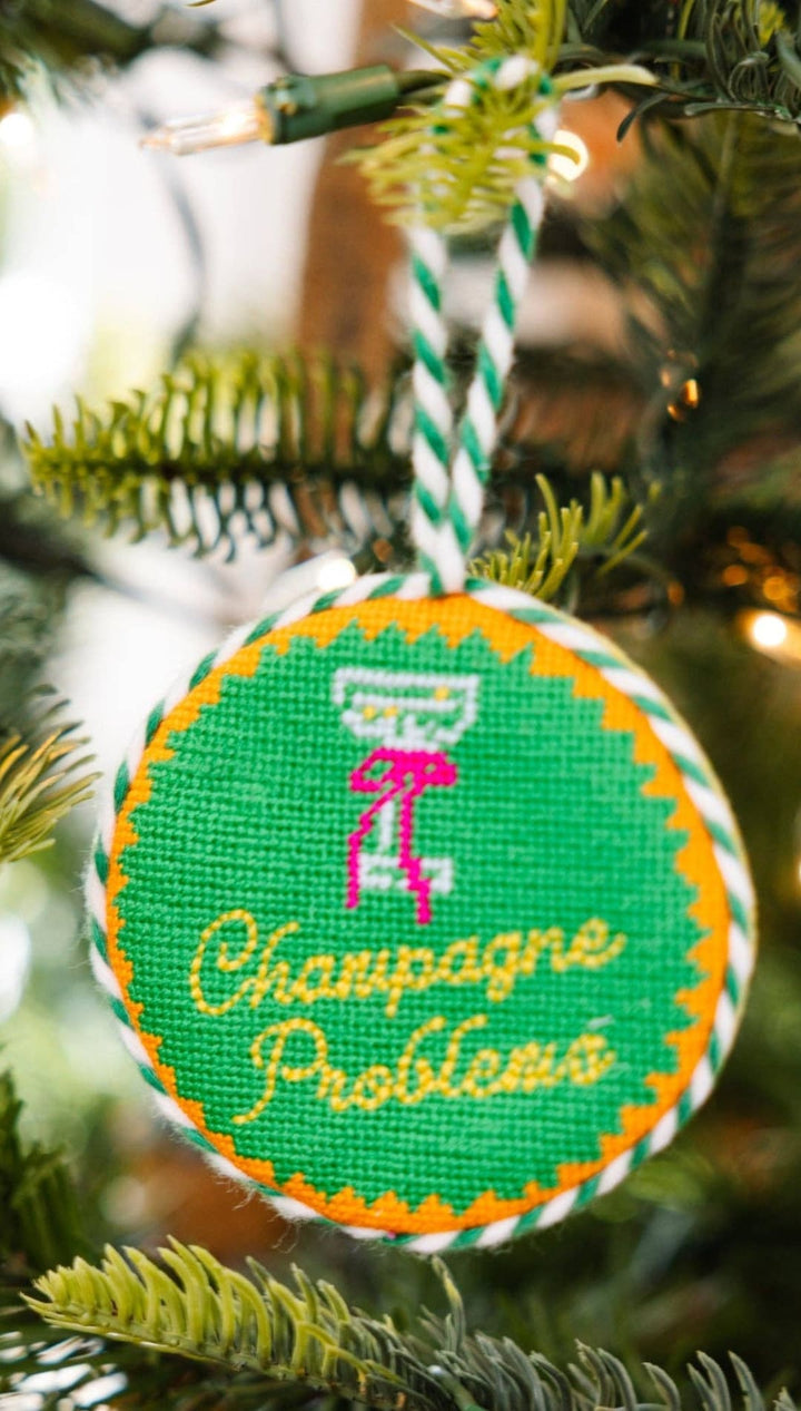 Green and yellow needlepoint ornament with champagne glass design, ’champagne problems’ text.