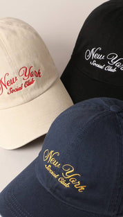 Three baseball caps in cream, black, and navy blue with ’new york social club’ embroidered in contrasting thread.