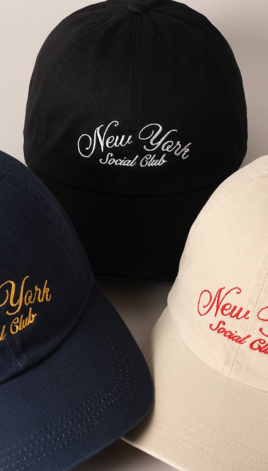 Three baseball caps in navy, black, and beige with ’new york social club’ embroidered in gold, white, or red thread.