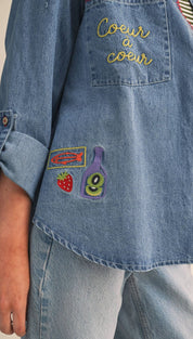 Blue denim shirt featuring embroidered patches with ’coeur à coeur’ in gold thread and colorful appliqués of a fish, strawberry, and bottle.