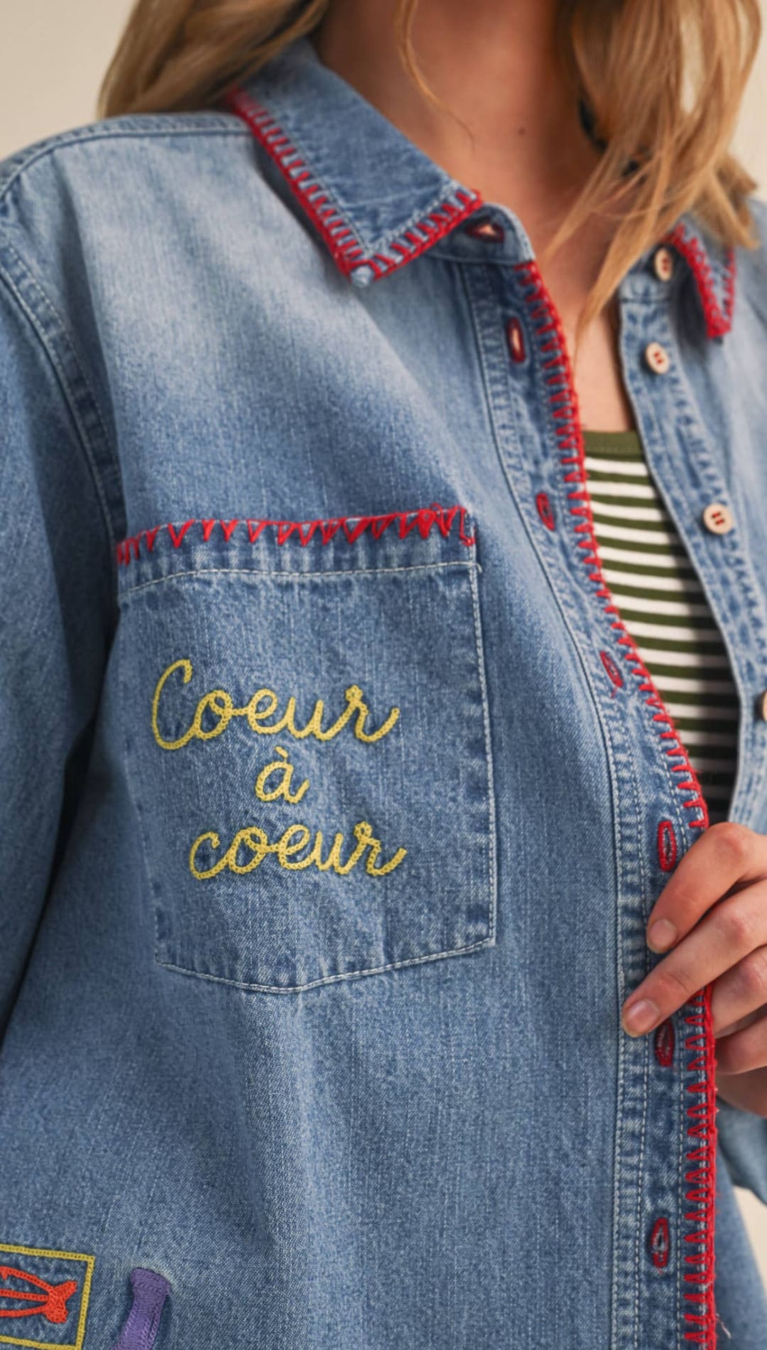 Blue denim shirt with red-stitched collar and pocket, featuring gold thread embroidery reading ’coeur à coeur’ on the chest.