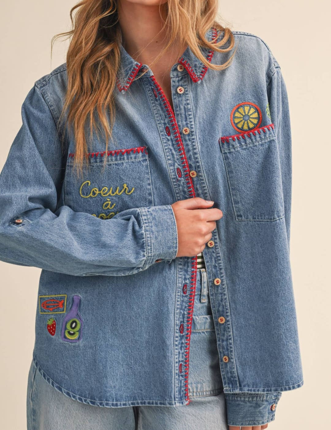 A light-wash denim shirt featuring colorful embroidered patches and red-trimmed pockets.