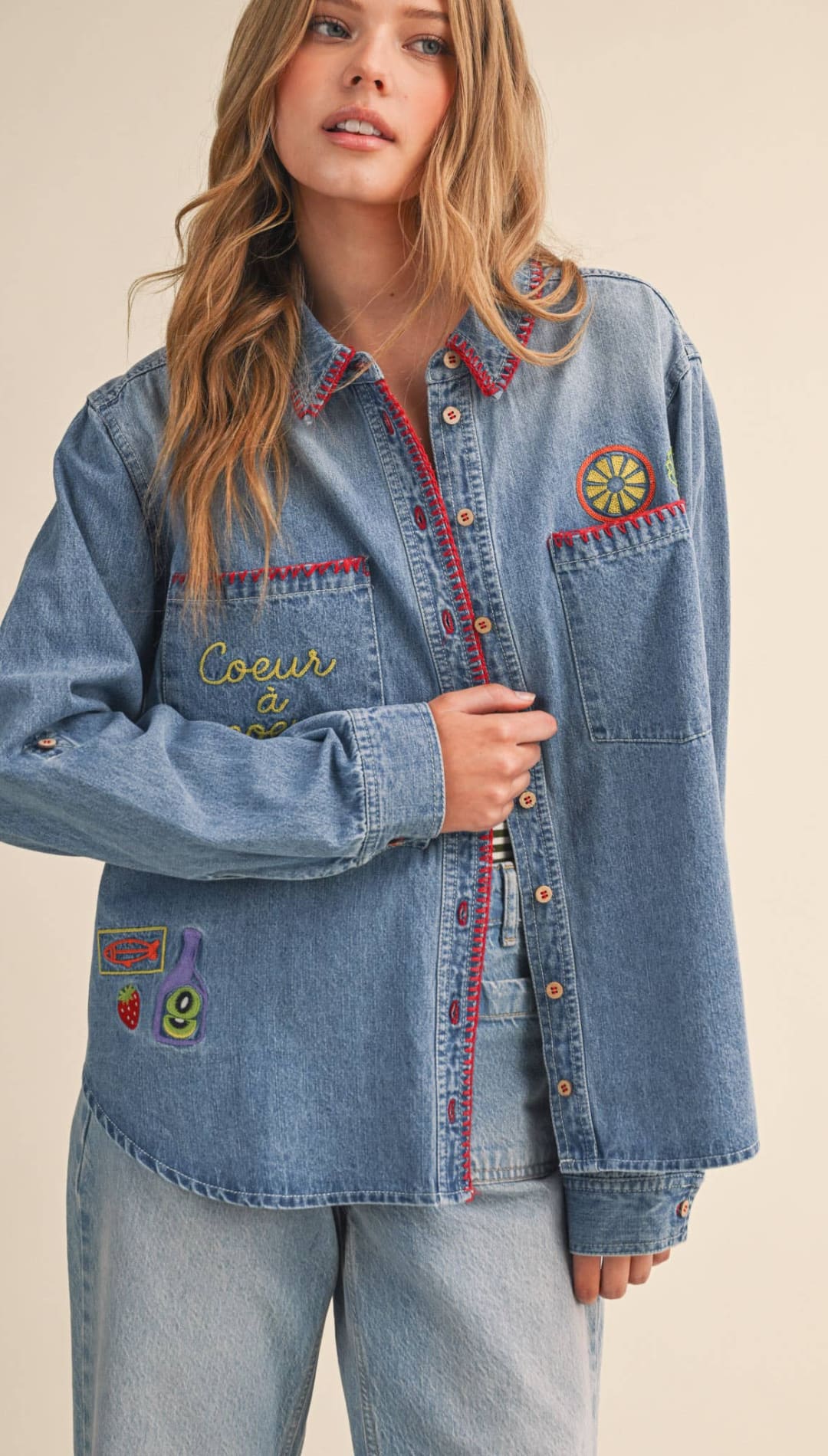 A light-wash denim shirt featuring colorful embroidered patches and red-trimmed pockets.