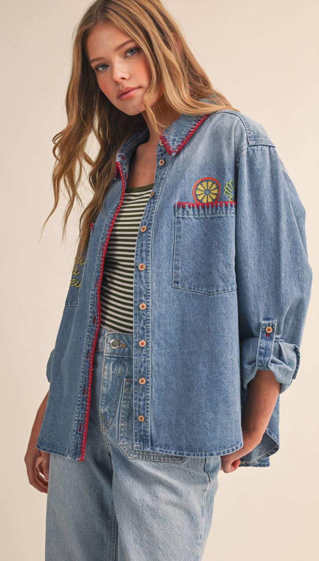 A medium-wash denim shirt with red-trimmed collar and cuffs, featuring a colorful embroidered sun patch on the chest.