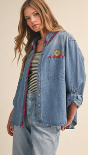 A medium-wash denim shirt with red-trimmed collar and cuffs, featuring a colorful embroidered sun patch on the chest.