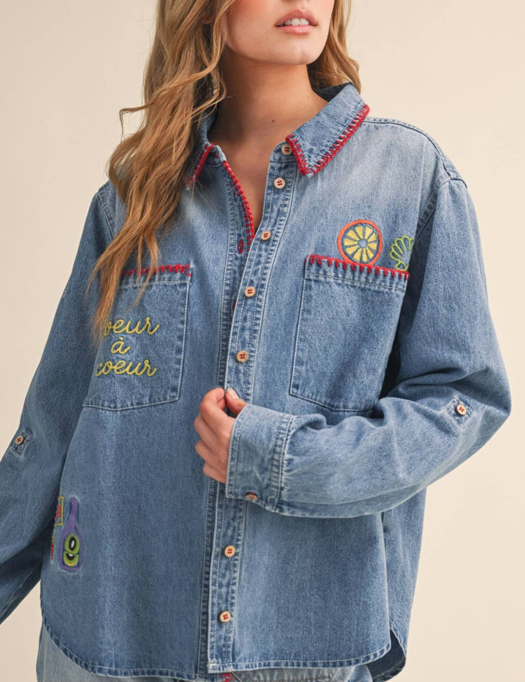 A light-wash denim button-up shirt featuring colorful embroidered patches and a red-trimmed collar.