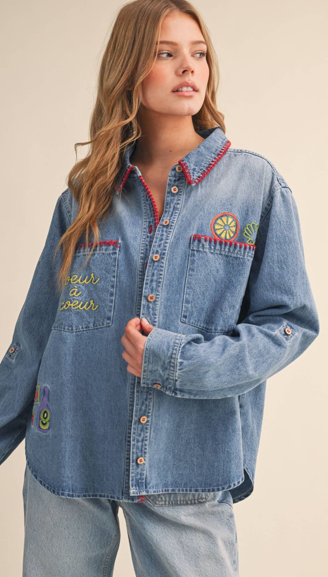A light-wash denim button-up shirt featuring colorful embroidered patches and a red-trimmed collar.