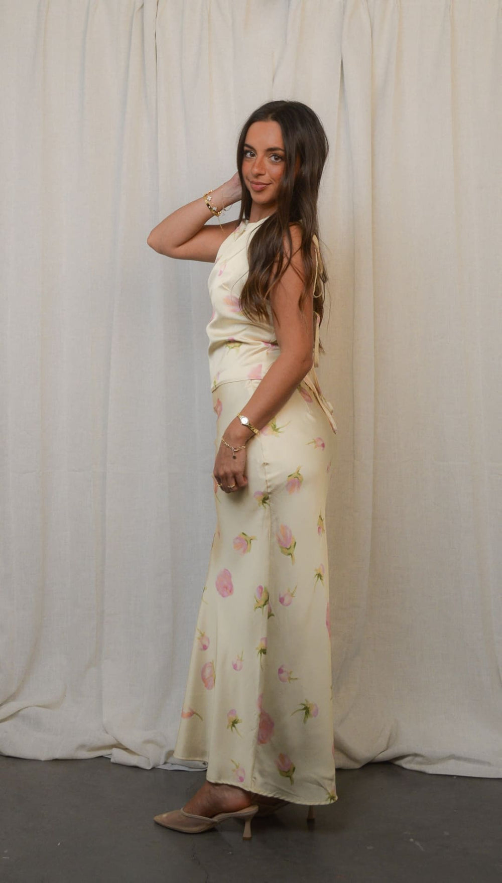 A woman modeling a long, off-white dress with a subtle pink floral pattern and a sleeveless top, paired with light brown high-heeled mules.