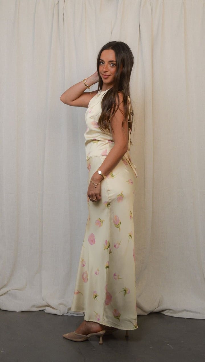 A long, light-colored dress with a subtle floral print, featuring a sleeveless top and a flowy skirt, paired with light-toned heels.