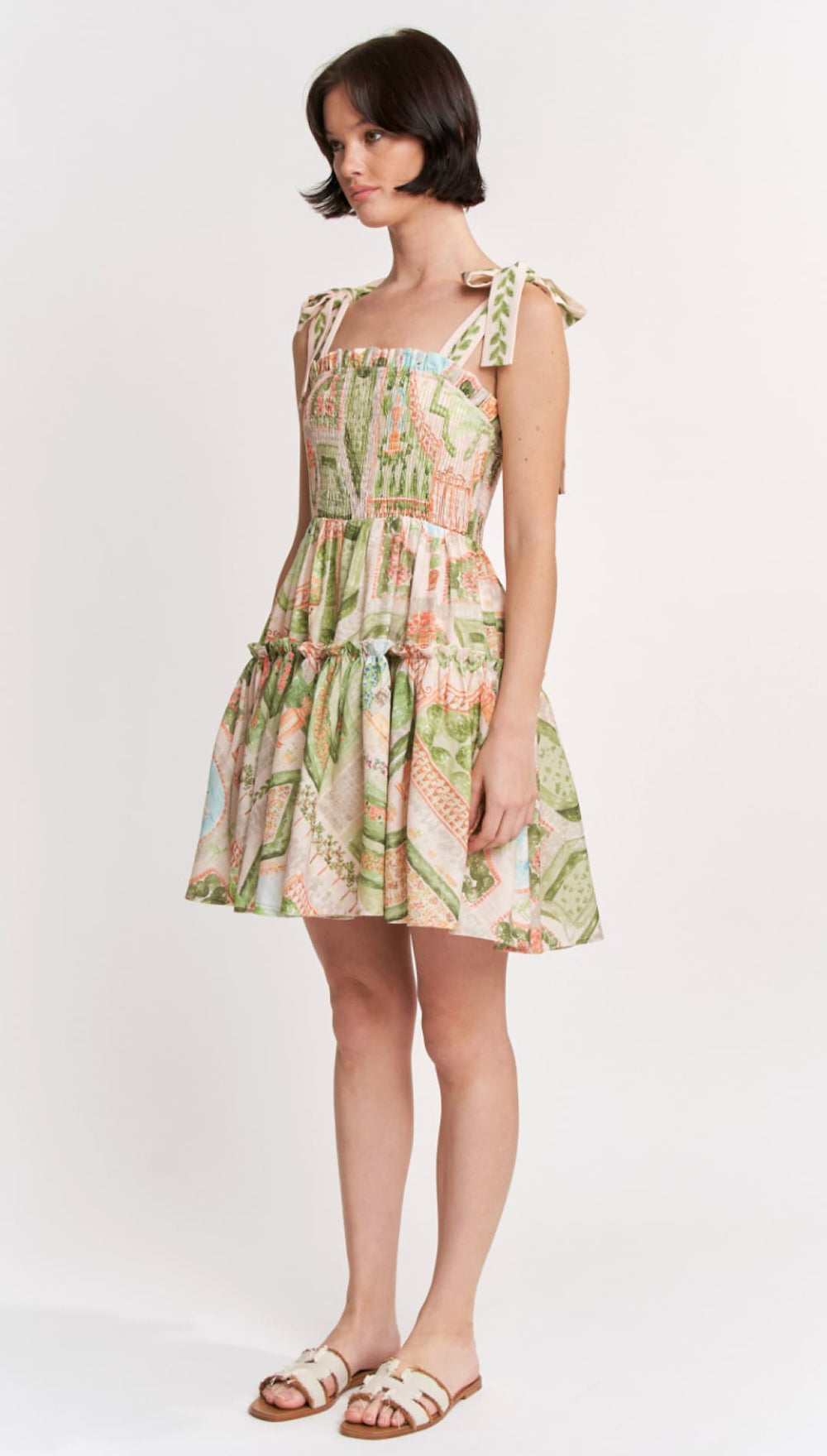 A woman wears a short, sleeveless dress with a square neckline and tied shoulder straps, featuring an intricate botanical print in soft greens, pinks, and cream on lightweight fabric.