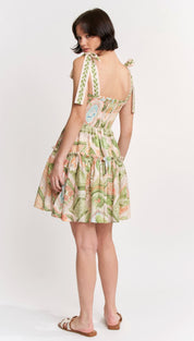 A woman wears a short, sleeveless dress with tie-back straps and an all-over botanical print in soft greens, pinks, and cream on a lightweight fabric.