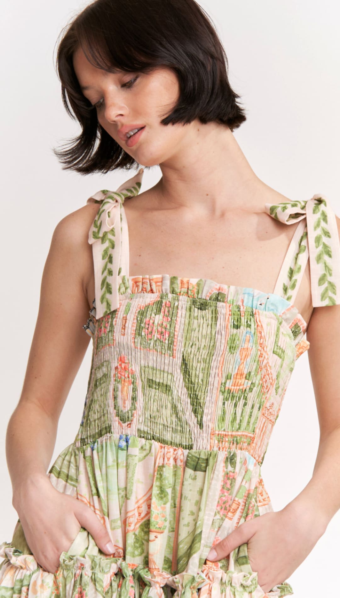 A woman wears a sleeveless, tiered dress with tie-back straps and an intricate botanical print in shades of green, peach, and cream.