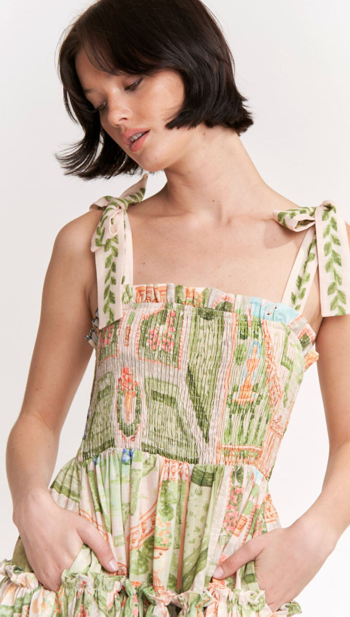 A woman wears a sleeveless, tiered dress with tie-back straps and an intricate botanical print in shades of green, peach, and cream.