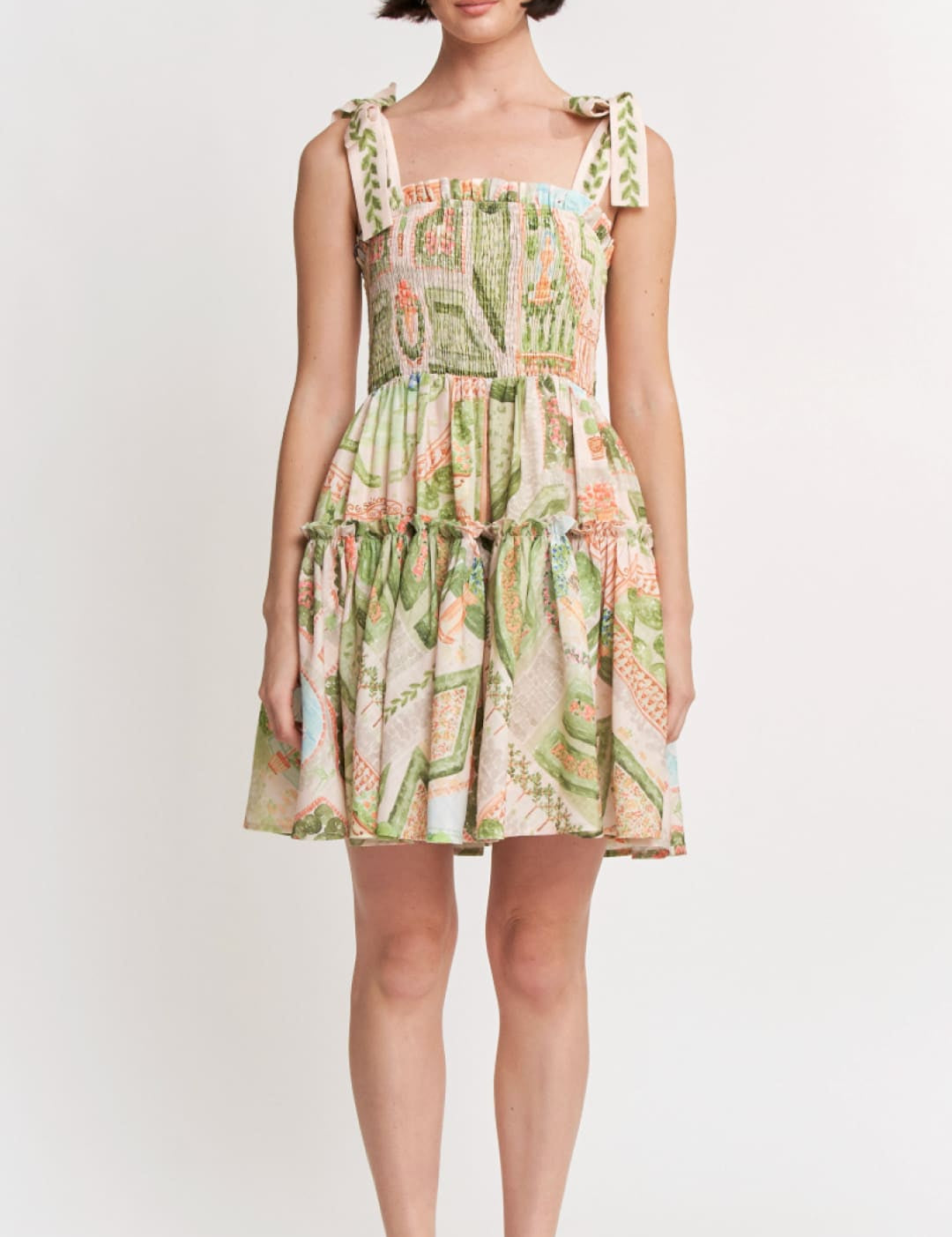 A woman wears a sleeveless, tiered mini dress with a vibrant botanical print in shades of green, pink, and beige.