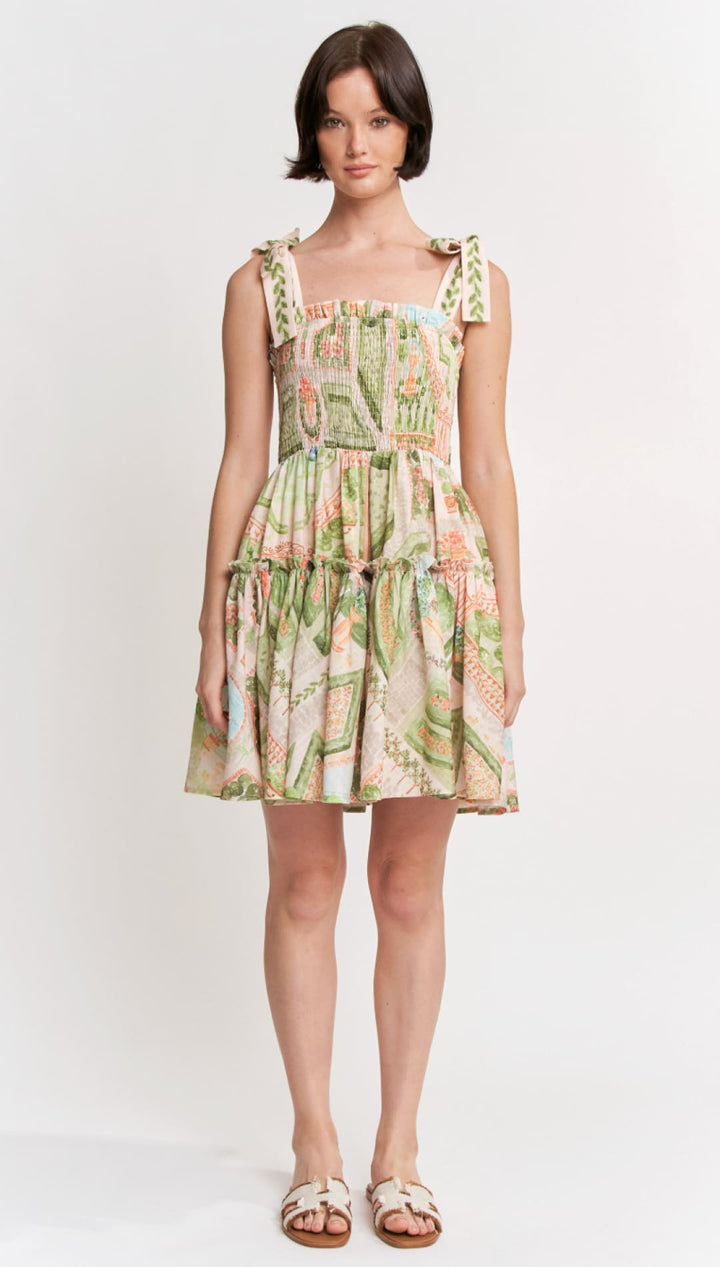 A woman wears a sleeveless, tiered mini dress with a vibrant botanical print in shades of green, pink, and beige.