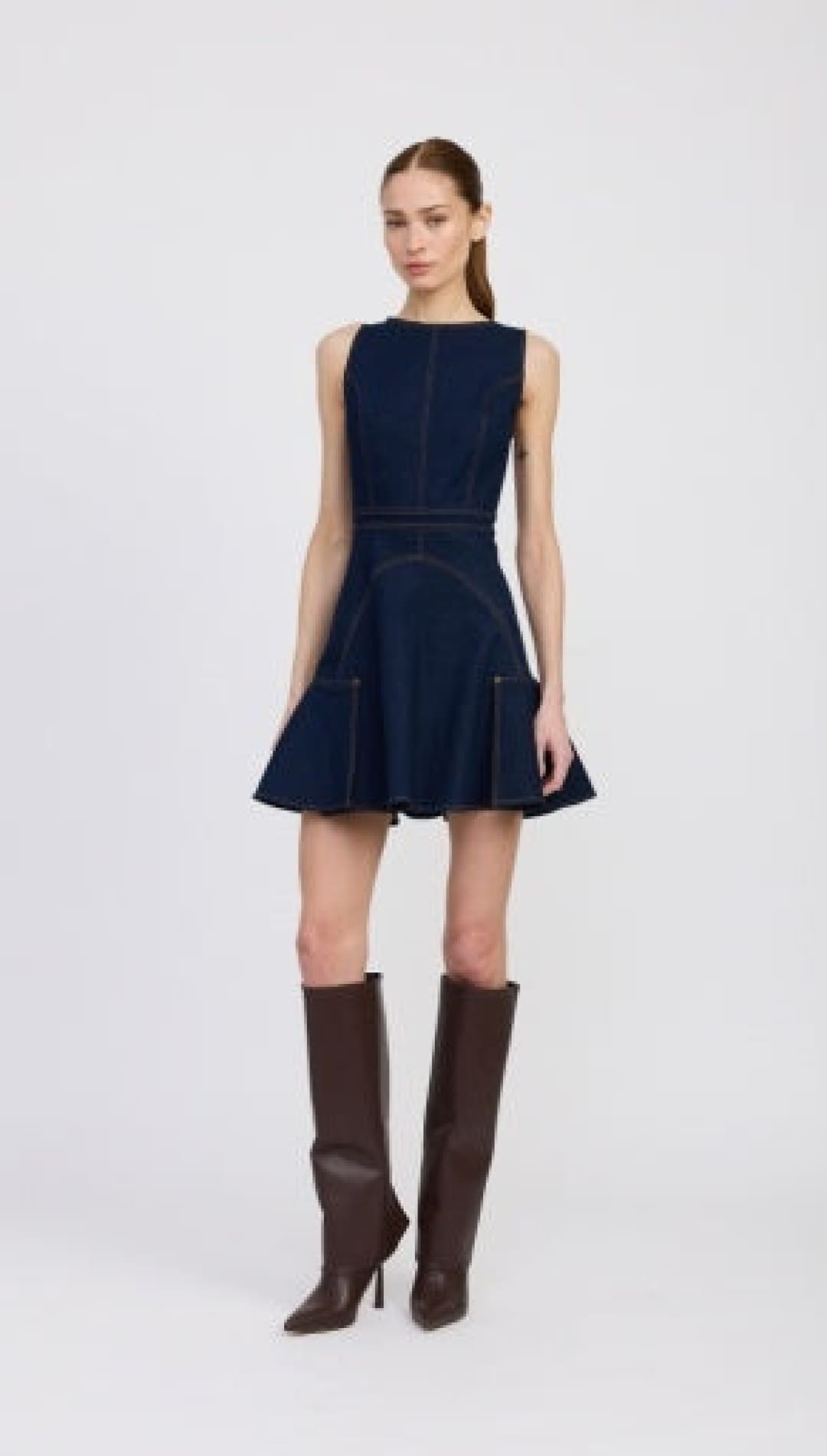 A dark blue denim sleeveless mini dress with a flared skirt and visible seam detailing, paired with dark brown knee-high heeled boots.