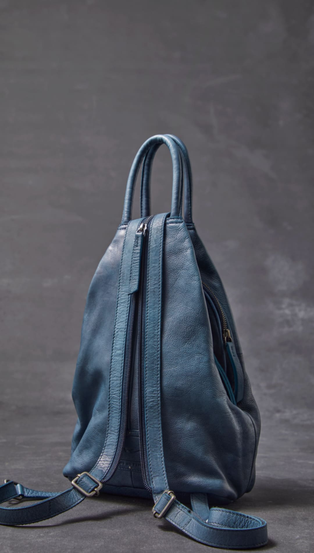 A soft, dusty blue leather backpack with a minimalist design and dual zippers.