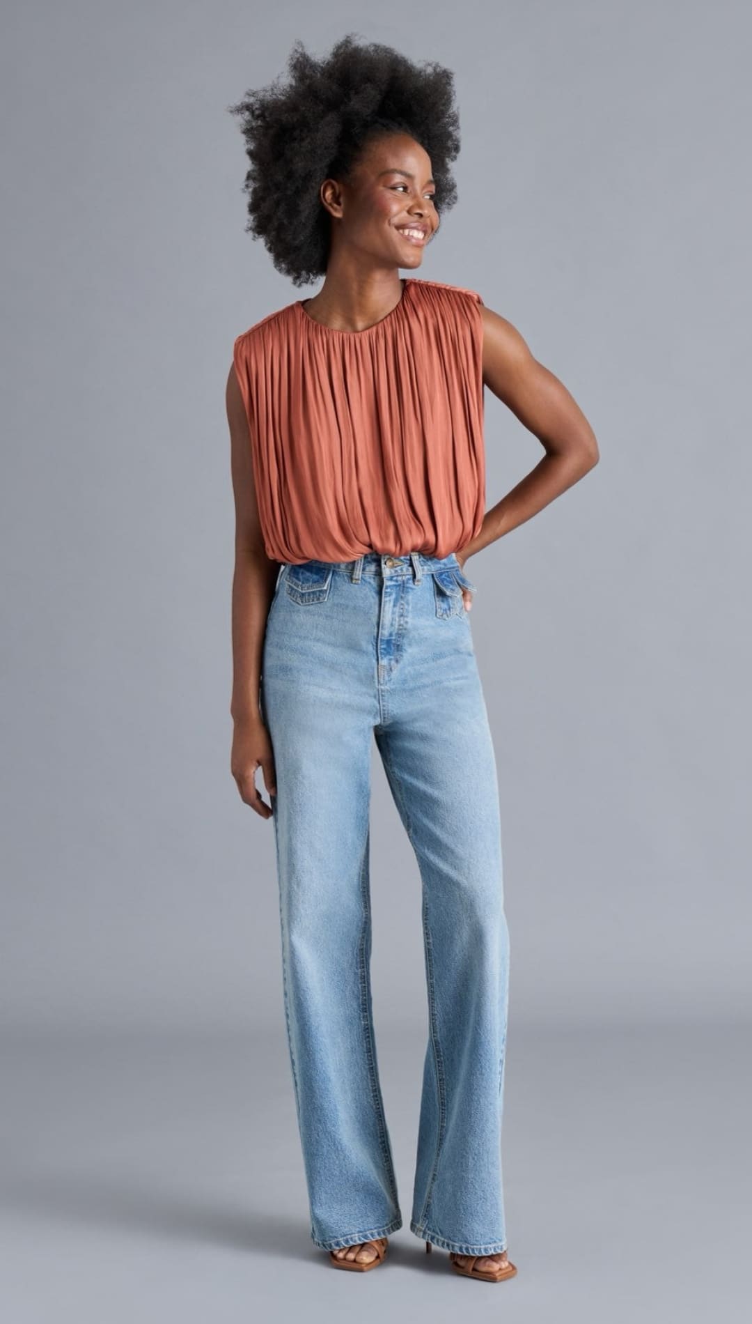 A woman wearing a pleated rust-colored sleeveless top and light wash wide-leg jeans.