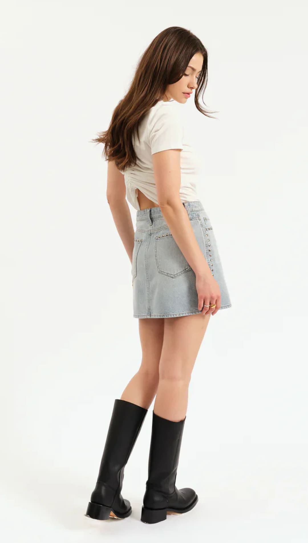 A young woman with long, dark hair wearing a white crop top, light blue denim mini skirt, and black knee-high boots.