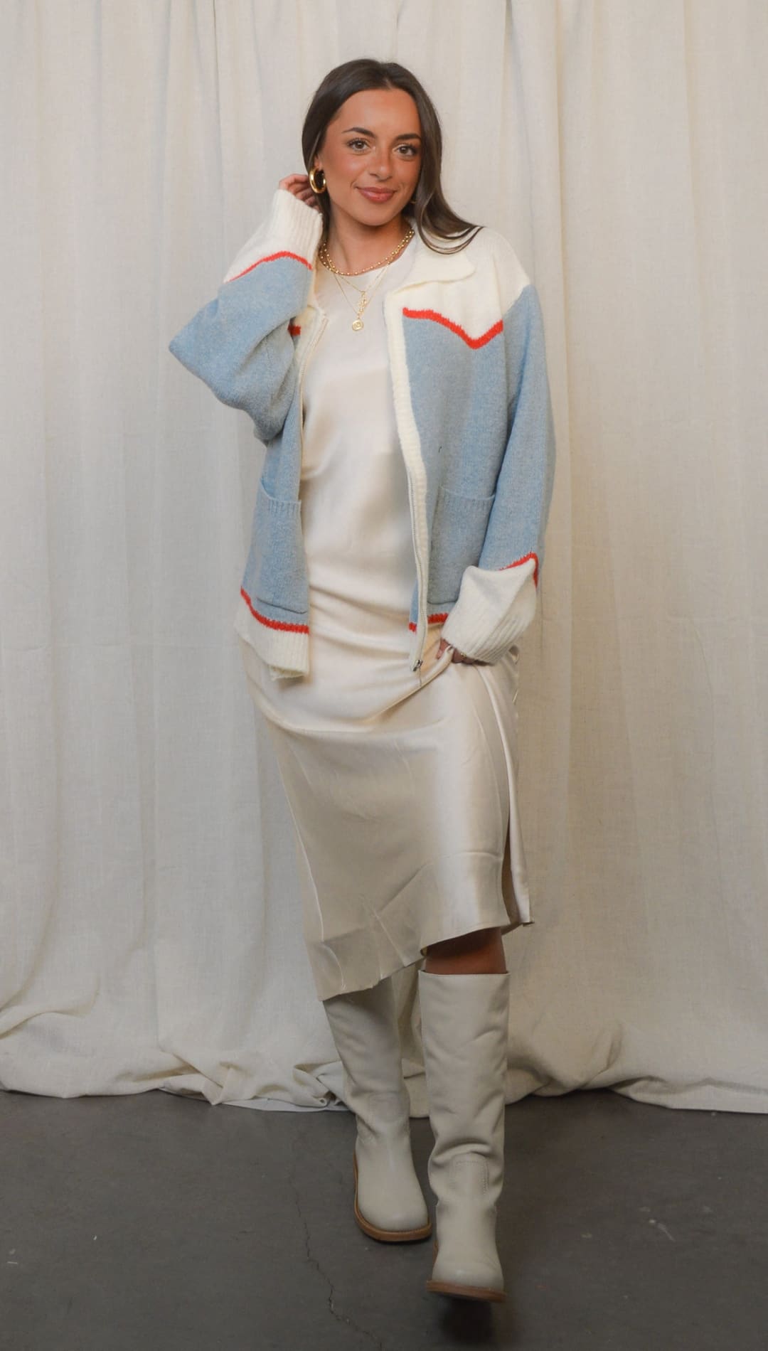 A stylish woman wearing a cream slip dress and a light blue and white cardigan with red trim, paired with cream knee-high boots.