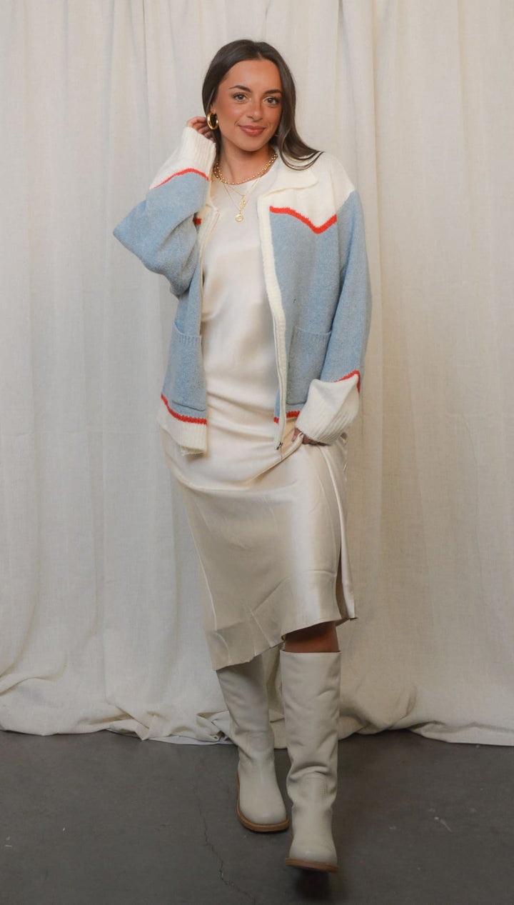 A stylish woman wearing a cream slip dress and a light blue and white cardigan with red trim, paired with cream knee-high boots.