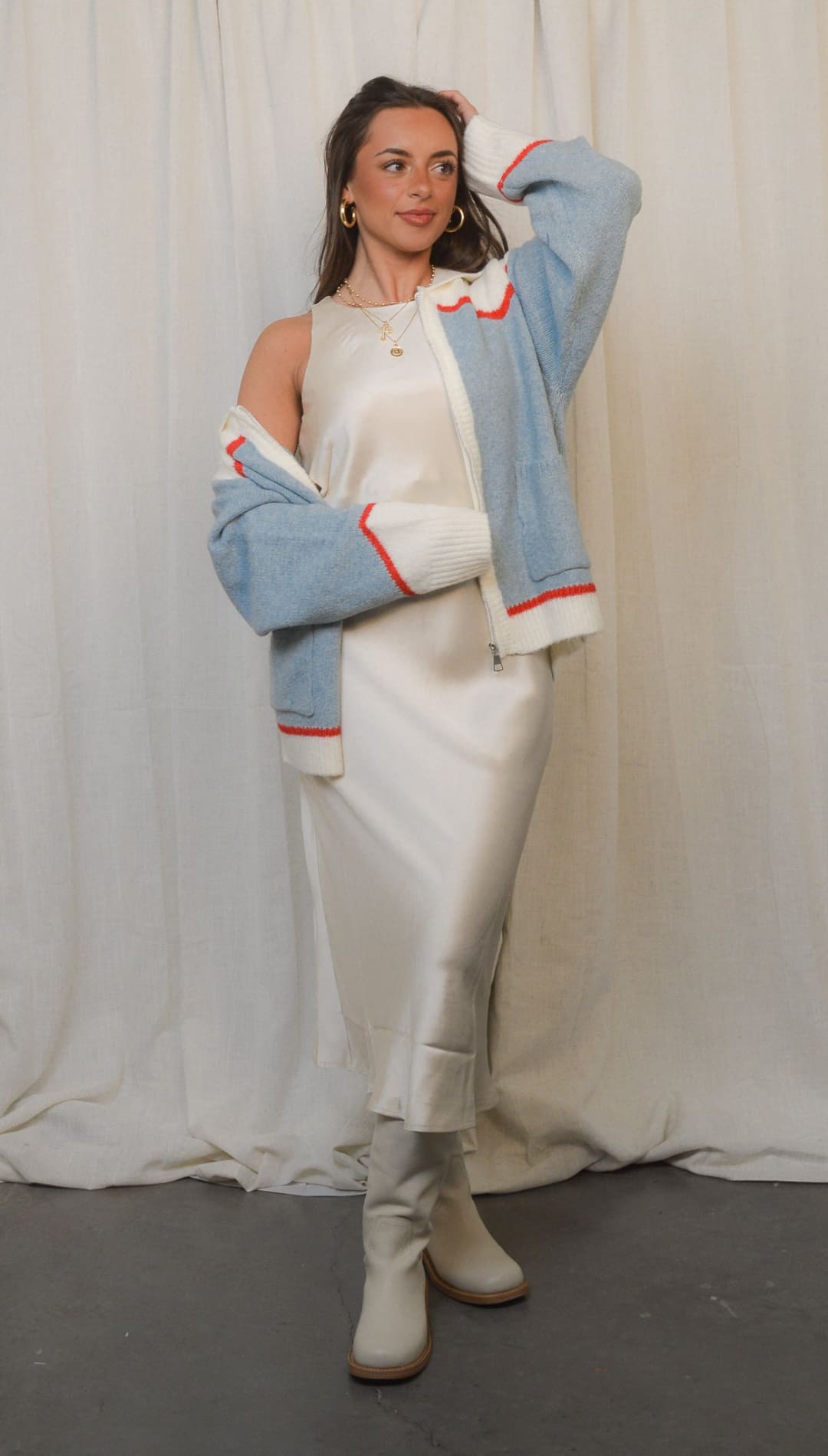 Woman in a sleeveless white dress, light blue and white color block jacket with red trim, and light-colored boots.