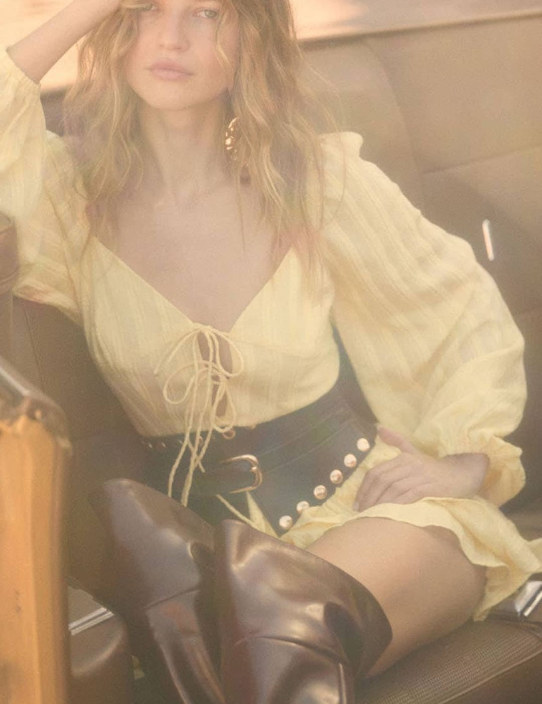A woman wearing a pale yellow, long-sleeved blouse with ruffled details and a lace-up front paired with black leather boots.