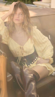 A woman wearing a pale yellow, long-sleeved blouse with ruffled details and a lace-up front paired with black leather boots.