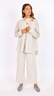 A woman wears a matching striped linen jacket and wide-leg pants in soft beige with thin vertical stripes, paired with woven blue flat shoes.