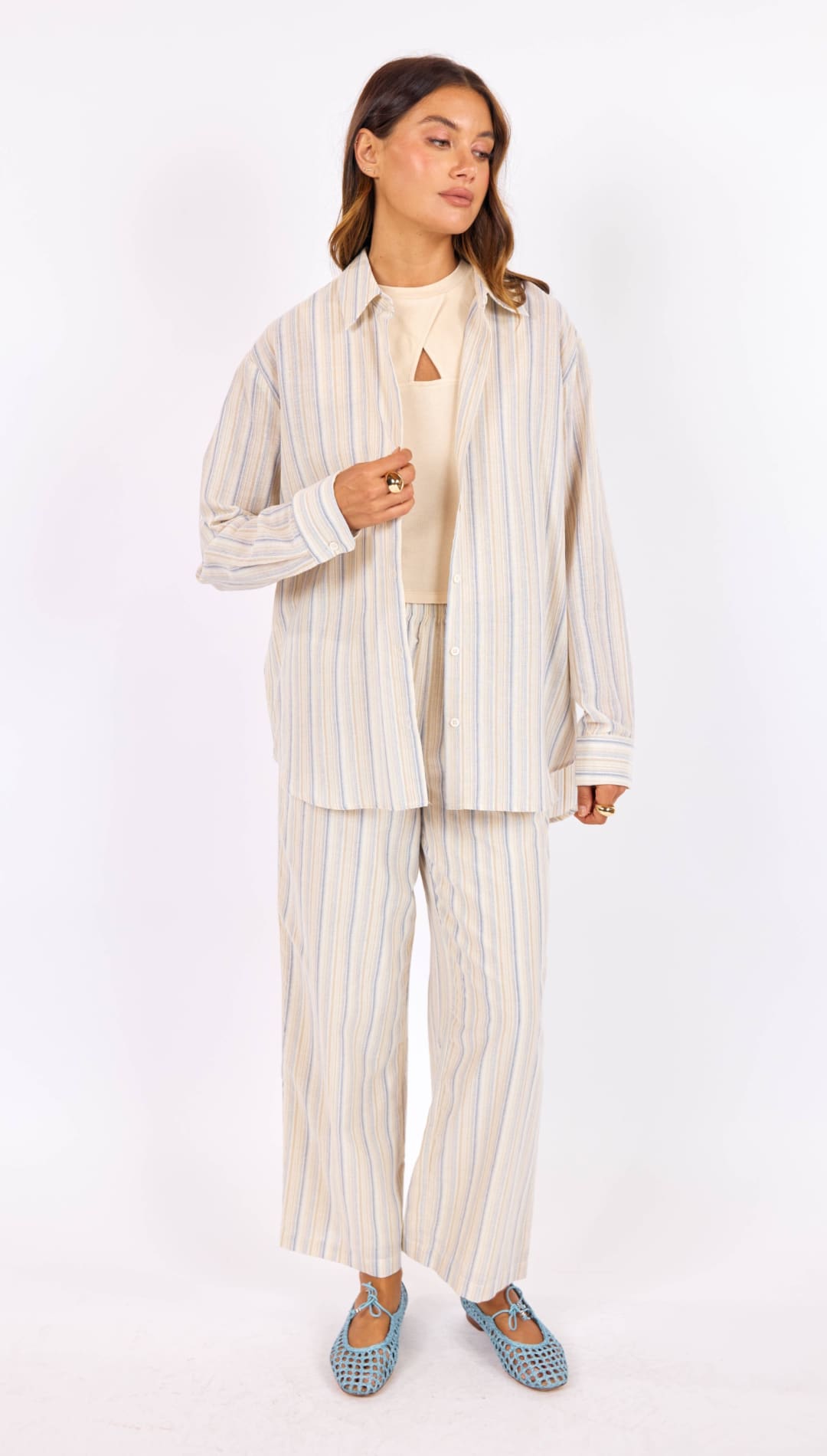 A woman wears a loose-fitting, vertically striped button-up shirt in soft beige and pale blue tones with matching wide-leg trousers.