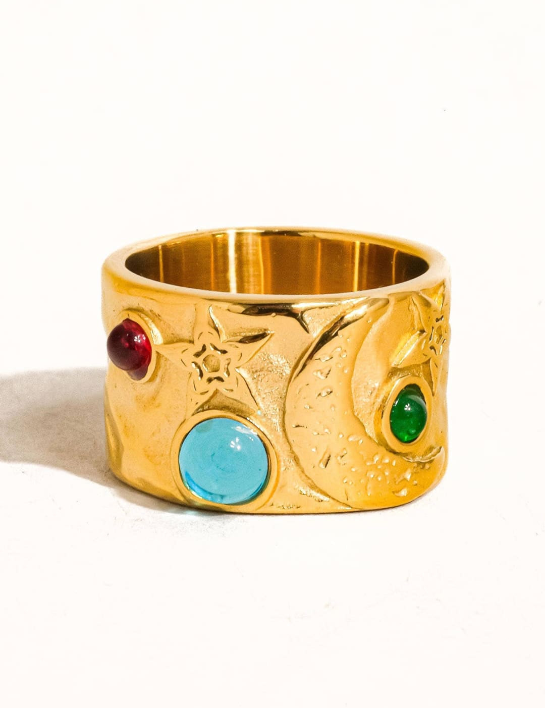 Gold-colored ring with a textured, wide band featuring three embedded gemstones — one red, one turquoise blue, and one green — set against an engraved starry design.