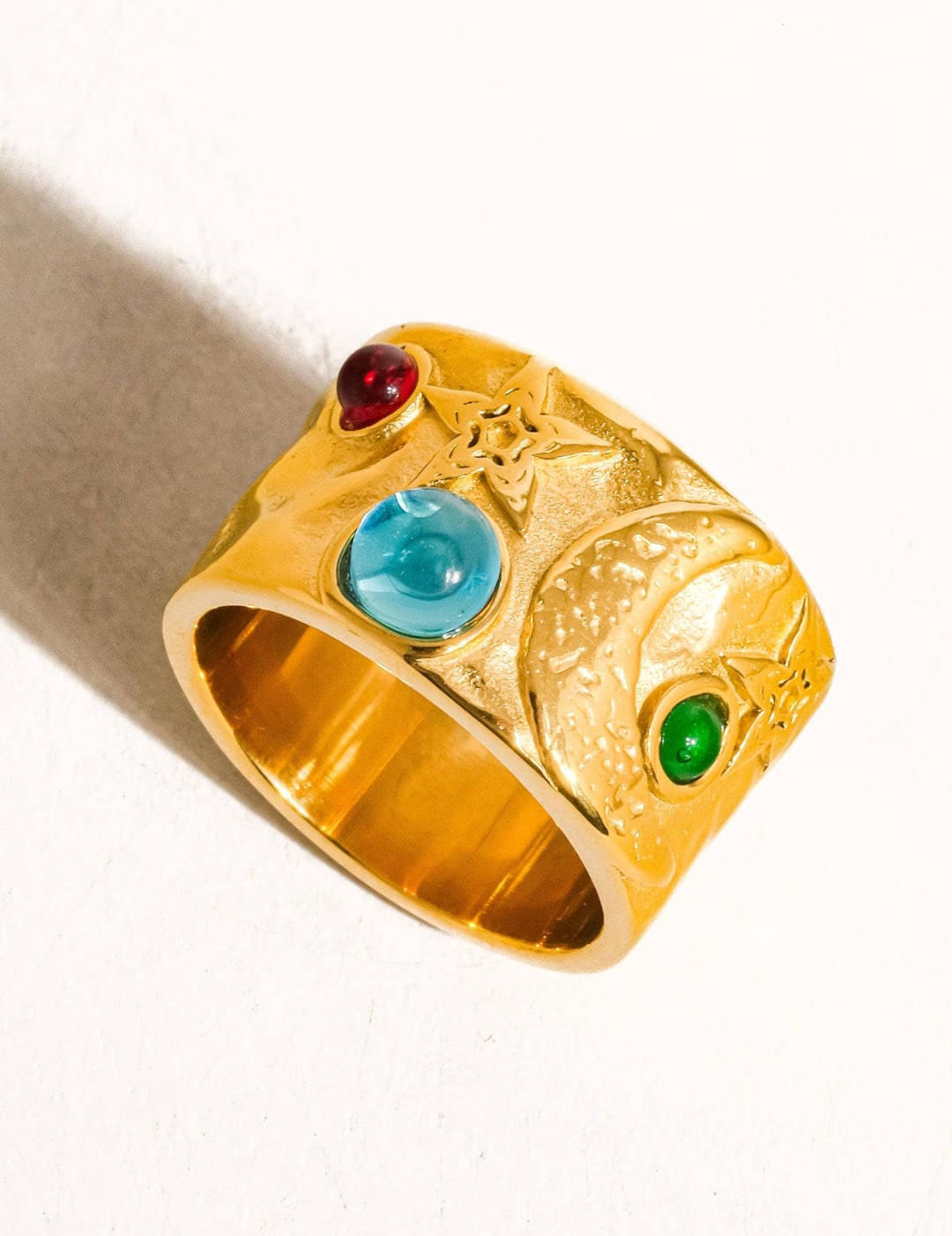 Gold-colored ring with a textured, sculpted design featuring three embedded gemstones: a red ruby, a blue sapphire, and a green emerald.
