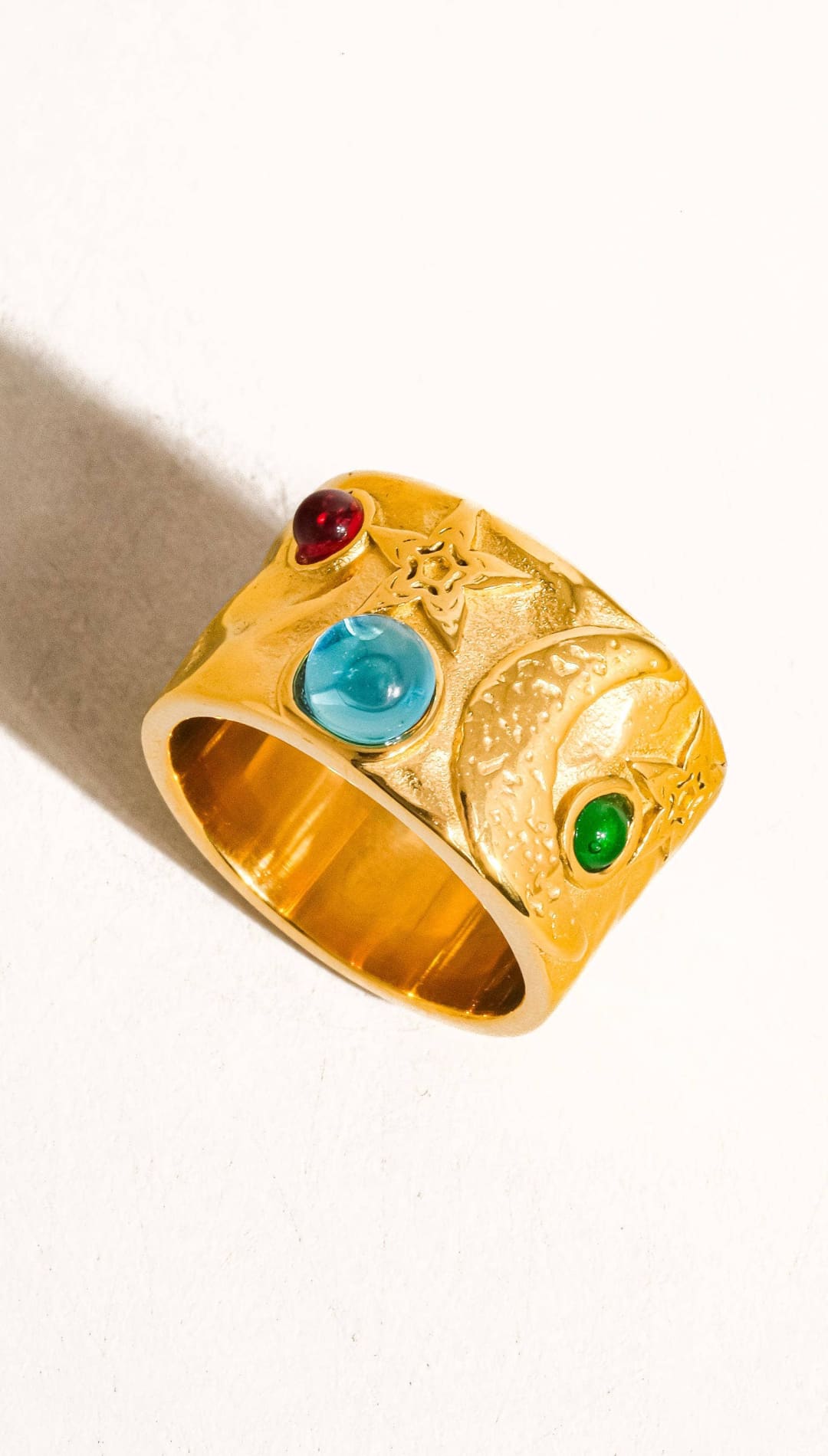 Gold-colored ring with a textured, sculpted design featuring three embedded gemstones: a red ruby, a blue sapphire, and a green emerald.