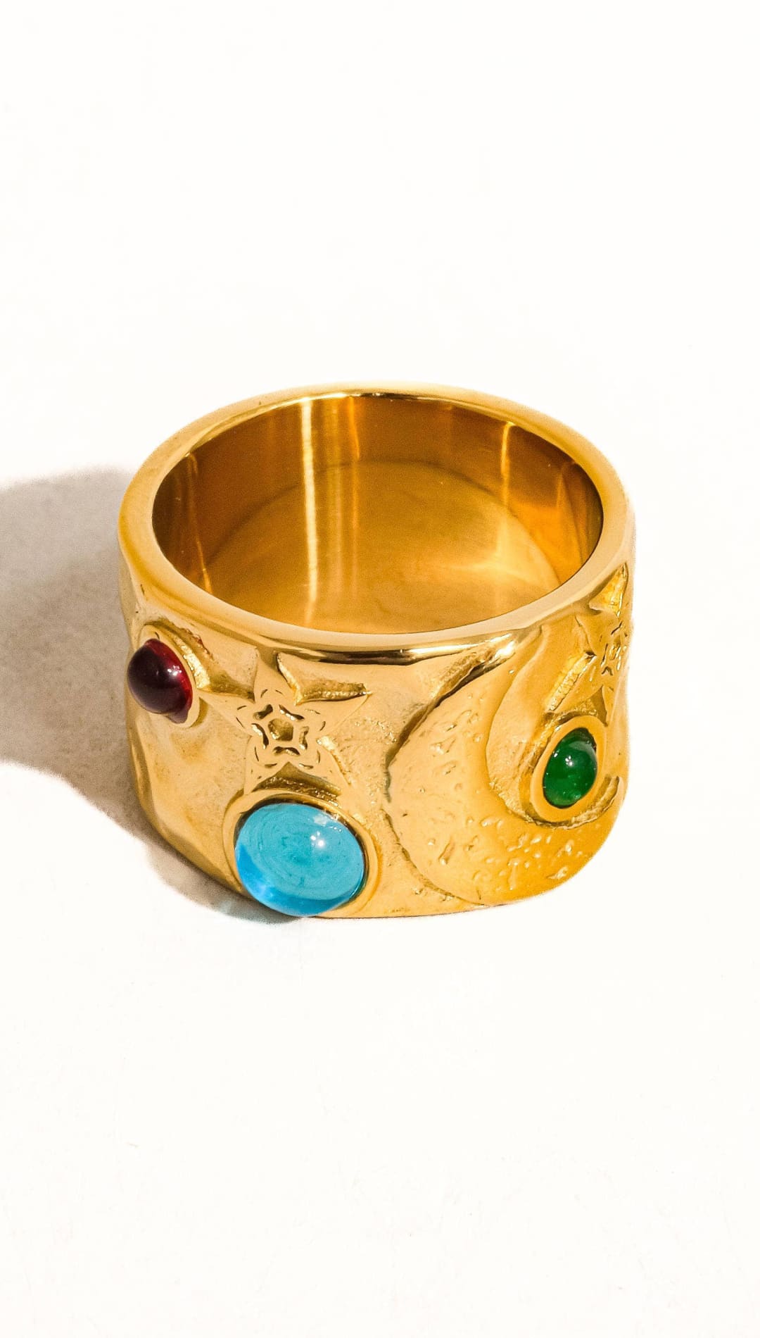Gold-colored, wide band ring featuring three prominent gemstones—red, green, and blue—and intricate engraved details resembling the infinity gauntlet.