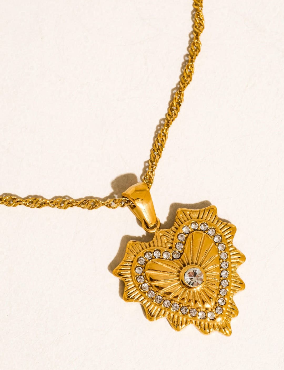 Gold heart-shaped pendant with a sunburst design and embedded crystals, hanging from a matching gold chain.
