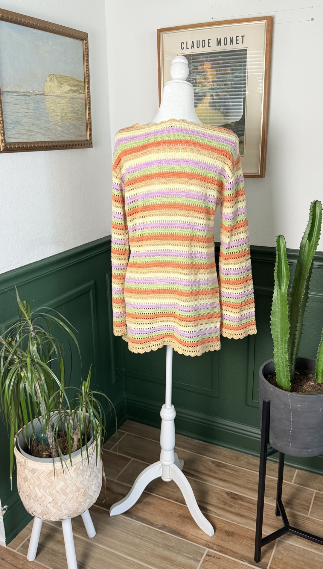A soft, hand-knitted sweater with horizontal stripes in pastel shades of peach, lavender, and cream hangs on a white mannequin.