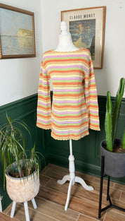 A vibrant, hand-knitted sweater in horizontal stripes of orange, peach, lavender, and cream, displayed on a white mannequin.