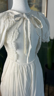 A delicate off-white lace-trimmed dress with a soft, flowing fabric and a charming bow at the neckline.