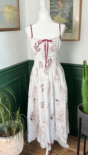 A white sundress with red embroidered botanical patterns and thin spaghetti straps, featuring a tied bow at the neckline.