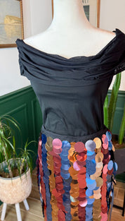 A black off-the-shoulder top with a ruched neckline and sheer lace sleeves, paired with a vibrant multicolored sequin skirt featuring vertical rows of iridescent discs in shades of pink, blue, orange, and gold.