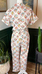 A vintage-style floral print jumpsuit in soft cream fabric, featuring short sleeves and a belted waist with rolled cuffs.