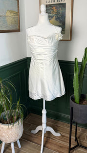 A white satin strapless dress with a ruched bodice and a small floral embellishment on the shoulder, displayed on a mannequin.